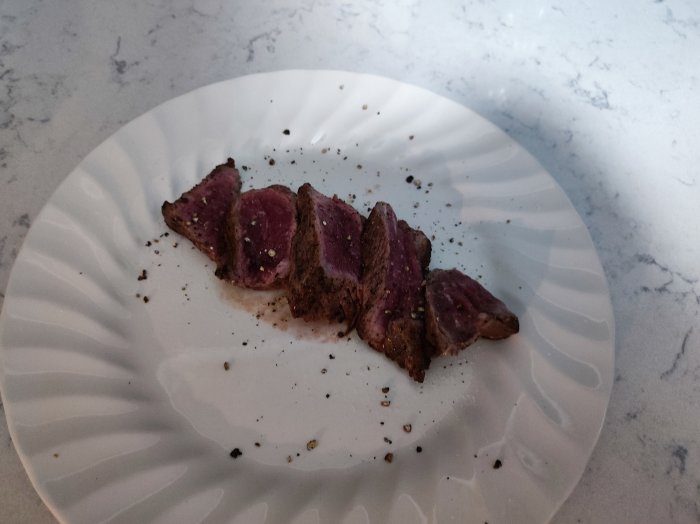 Skivat, rosa kött på vit tallrik, svartpeppar, grå marmorbänk.