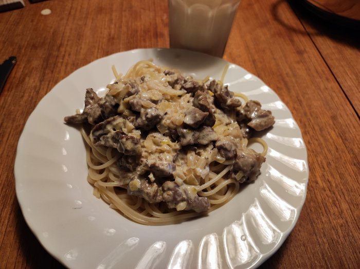 Ett vitt matfat med spagetti och stroganoff, mjölkglas i bakgrunden, träbord.