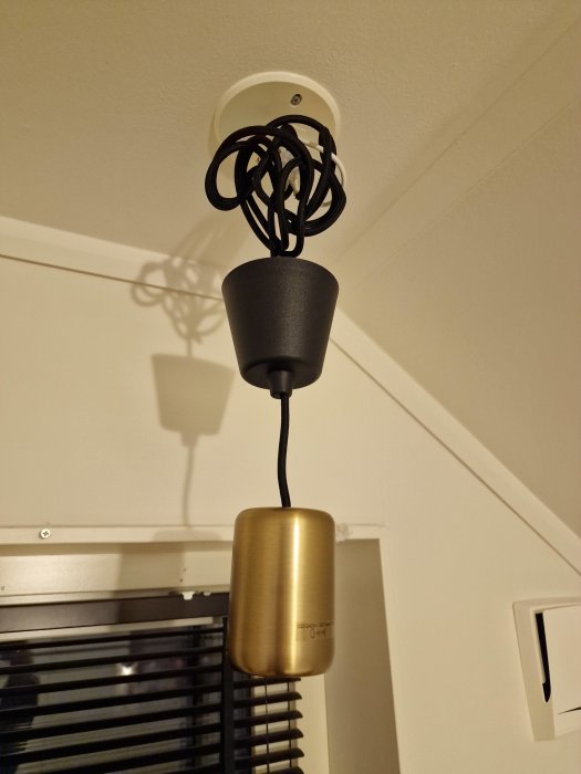 Lamphållare med trasslig kabel, guldskärma, mot vit takyta och vägg, skuggspel.