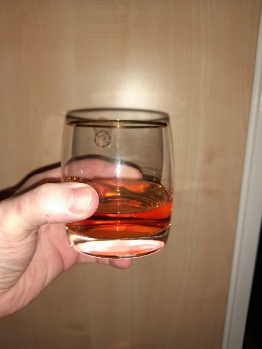 Hand håller ett halvfullt whiskyglas mot en enfärgad bakgrund.