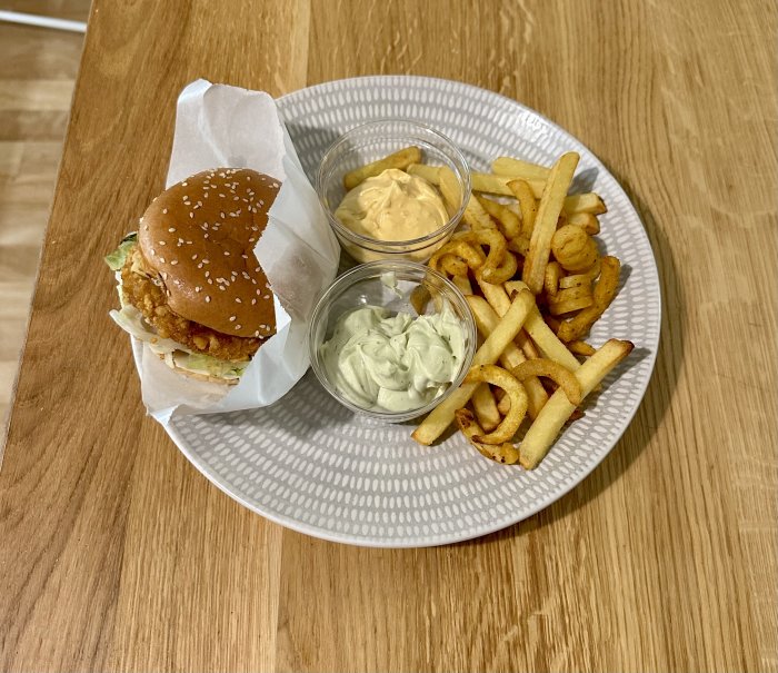 En hamburgare med sesamfrön, pommes frites, curly fries och två rör med sås på ett mönstrat fat.