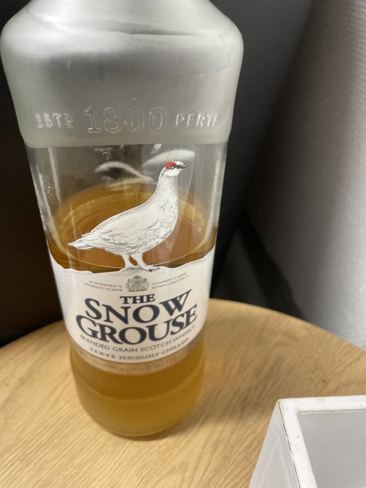 Flaska med The Snow Grouse whisky, förmodligen slatt kvar, mot bakgrund av trämöbel och grå väska.