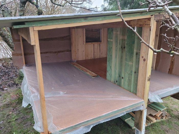 Träkonstruktion, halvfärdigt hus eller skjul, öppen vägg, bubbelplast på marken, träd i bakgrunden.