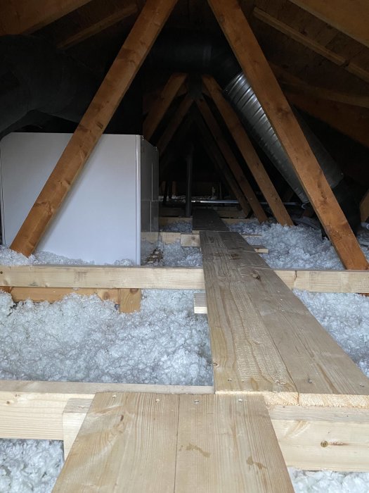 Vind med isolering, träbjälkar, planka gångväg och ventilationstrumma. Loftutrymme, obebott och konstruktionsdetaljer synliga.