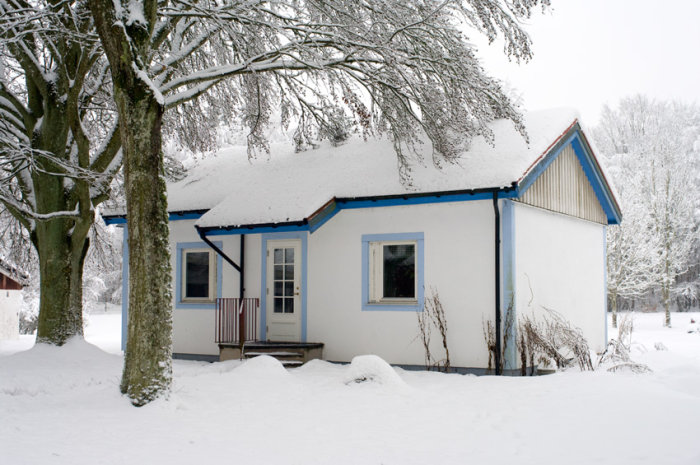 Ett vitt hus med blå detaljer omringat av snö och träd i vinterlandskap.