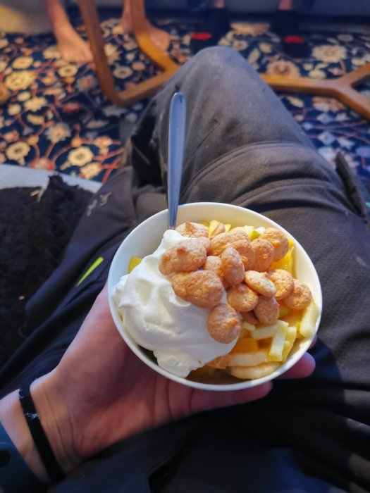 Person håller i skål med dessert, sittandes, mönstrad matta, trästol, barfota person i bakgrunden.