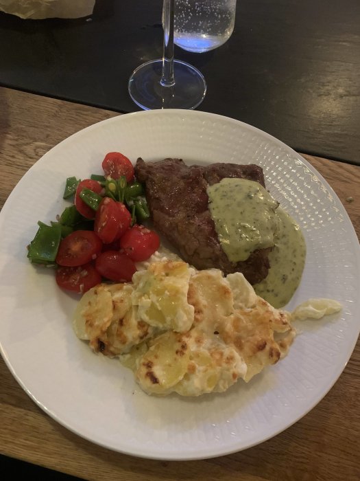 En måltid med biff, gratinerat potatismos, tomatsallad och Béarnaisesås, serverad på ett vitt fat.