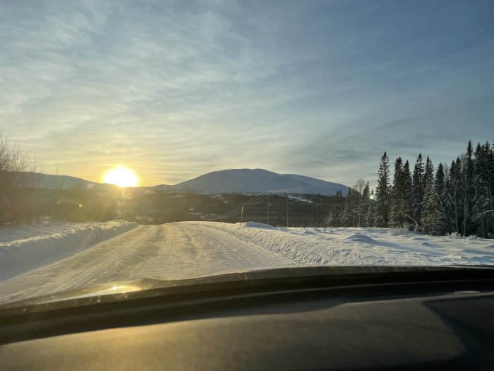 Solnedgång över snötäckt landskap, sett från bil, med träd och berg i bakgrunden.