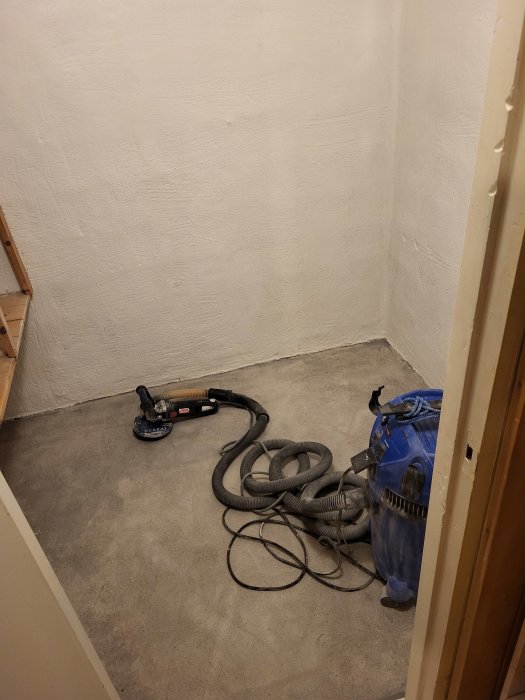 Tomrum med slipmaskin, dammsugare och sladdar på ett betonggolv, möjlig renovering pågår.