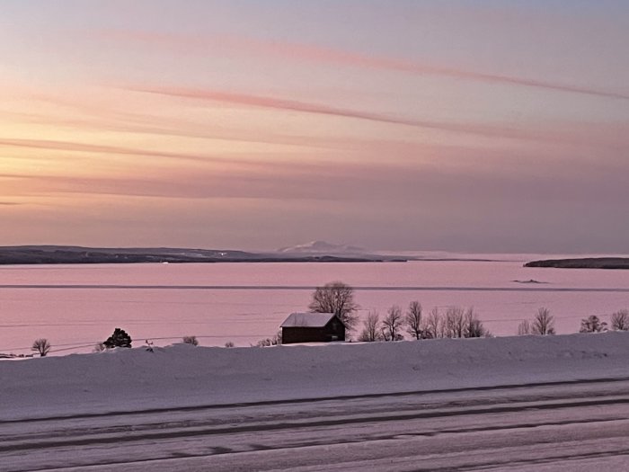 Vinterlandskap vid solnedgång med snötäckt mark, en ensam stuga och rosa himmel över isbelagt vatten.