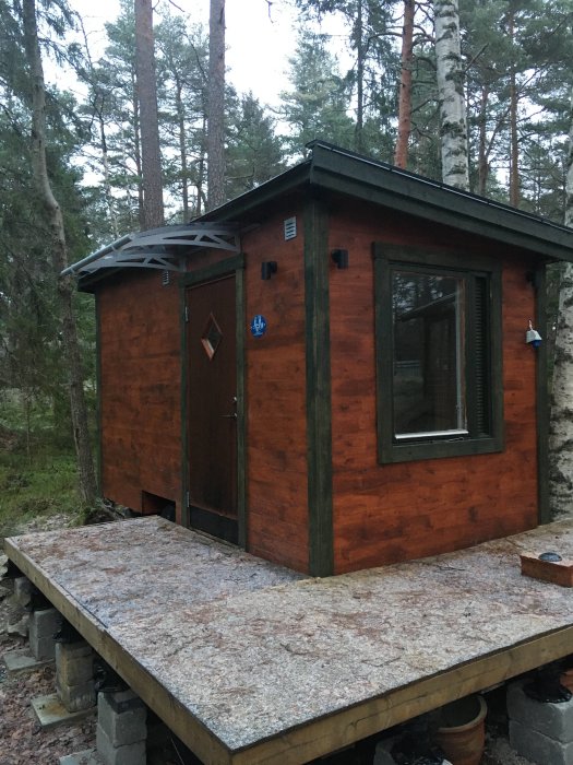 Träröd stuga i skogen med dörr, fönster, och plattform. Utomhus, grått ljus, tallar i bakgrunden.