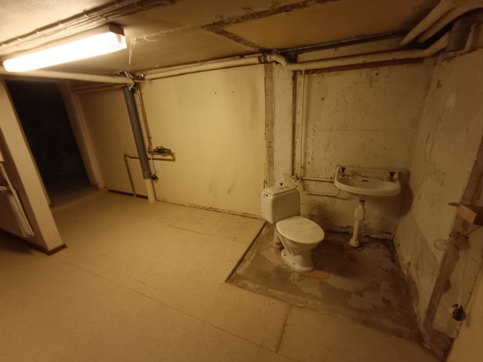 Oinredd källarlokal med toalett och handfat, belysning, rör på väggarna, behov av renovering.