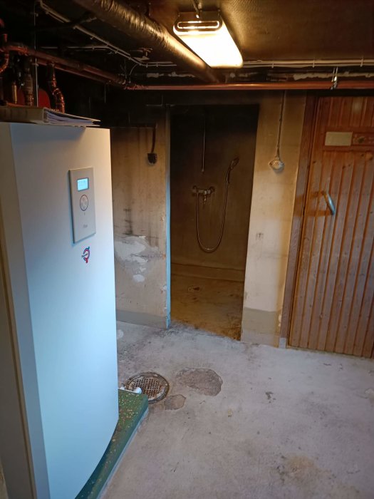 Oinredd källare med synliga rör, värmepanna, duschutrymme, betonggolv och träpaneler. Under renovering eller förvaring.