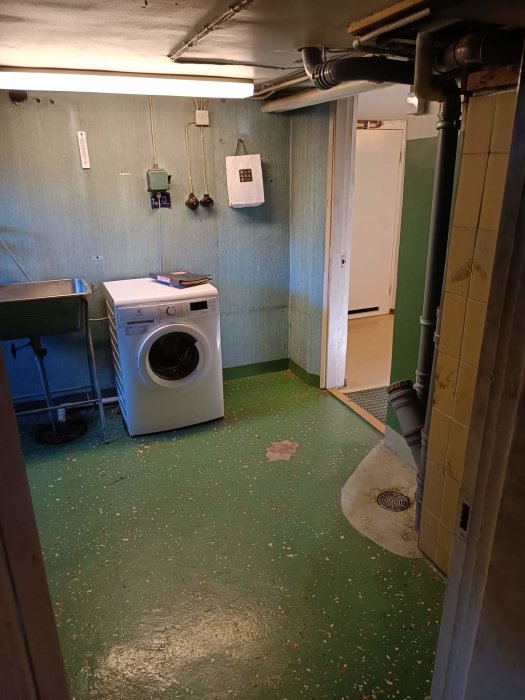 Tvättstuga med tvättmaskin, grön specklad golv, blå väggar, rör och bokningschema på väggen.