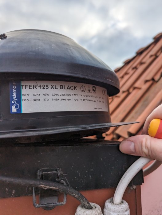 Elektrisk anordning med etikett, kablar, på tak, delvis hand syns, skymning eller molnig himmel i bakgrunden.