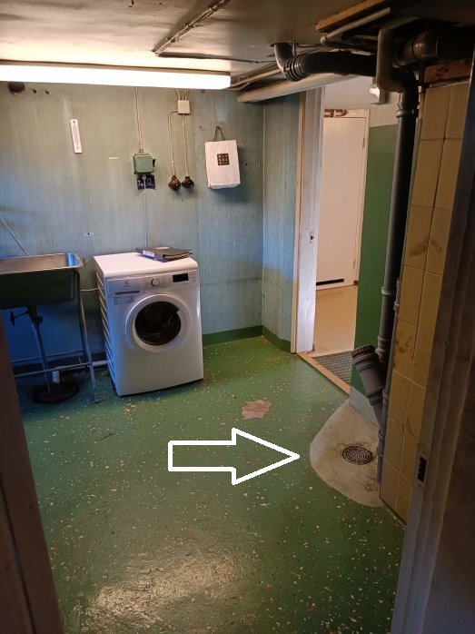 Källarlokal med tvättmaskin, grön golv, vit pil, rör i taket.