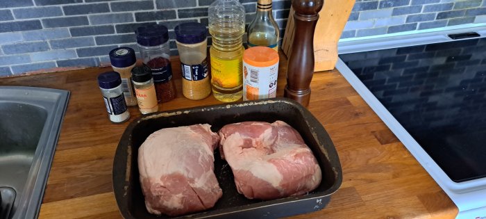 Två råa köttstycken i ugnsform, kryddor och oljeflaskor på köksbänk.