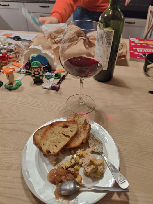Vin och mat på tallrik, person i bakgrunden, leksaker och oordning på köksbordet.