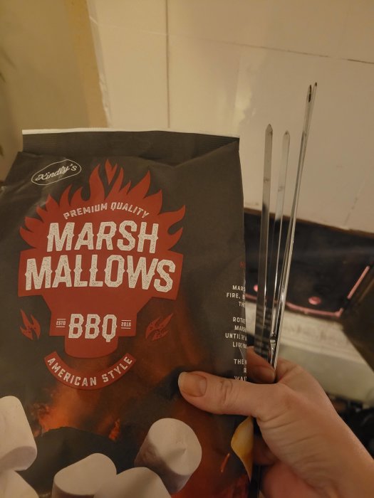 Förpackning med grillade marshmallows, hand håller, grillspett i bakgrunden, kakelvägg, inomhus, fritid, rostning.