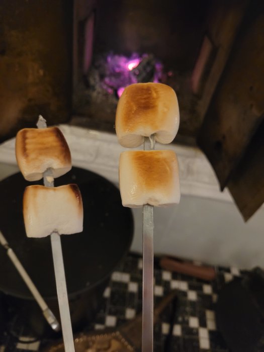 Fyra rostade marshmallows på spett framför en öppen spis med glödande kol.