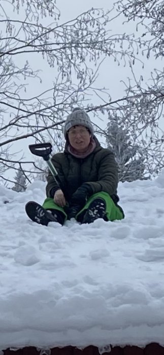 Person sittandes på snötäckt tak med snöskyffel, vinterkläder, träd och grå himmel i bakgrunden.