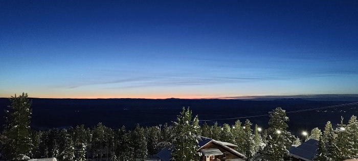 Vinterlandskap vid skymning med snöklädda träd, en stuga och en gradvis mörknande himmel.