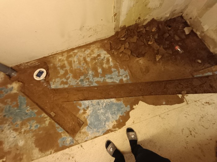 Förfallna byggnadsdetaljer, rör, sprucken färg, skräp, person i tofflor står och tittar ner.