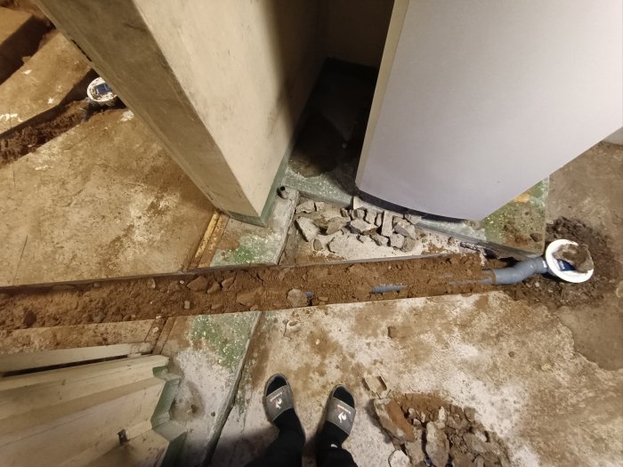 En rörledning under golvkonstruktionen, grus, betongrester, skor i förgrunden, byggarbetsplats.