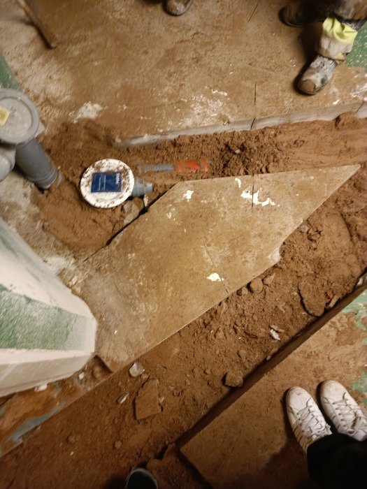 Uppbruten golv, rörinstallation, skor i förgrunden, inomhusarbetsplats med synligt smuts och stenmaterial.