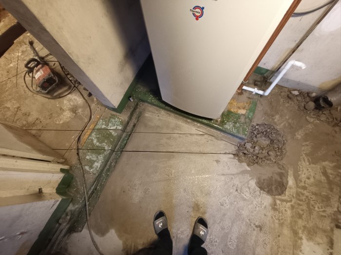 Oorganiserat utrymme med varmvattenberedare, kablar på golv, nedsmutsad betongyta och fötter i förgrunden.