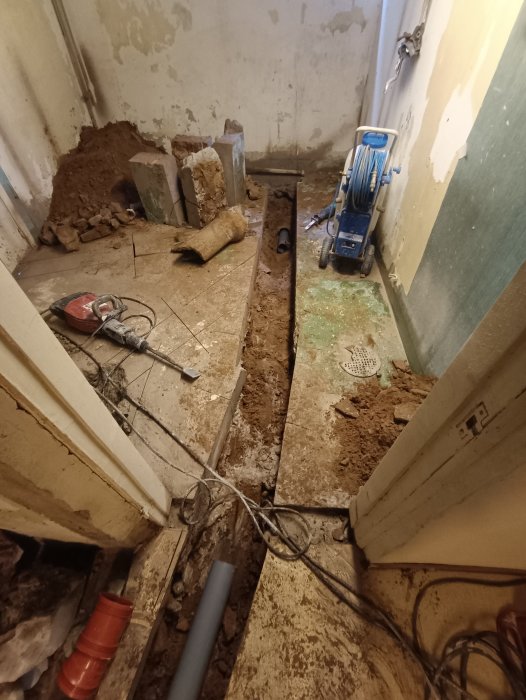 Renoveringsarbete, uppgrävd golv, synliga rör, byggavfall, sladdar, verktygsmaskin, skadad vägg, byggnadsplats inomhus.