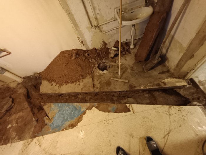 Ett skadat golv med spilld jord, rör, en handfat och någons fötter synliga; behov av renovering eller reparation.