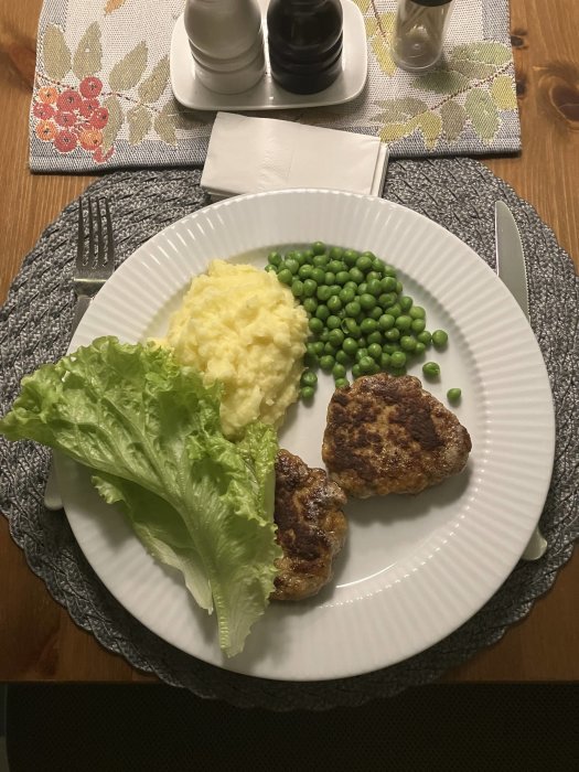 Middagstallrik med köttbullar, potatismos, gröna ärtor och salladsblad på ett träbord.