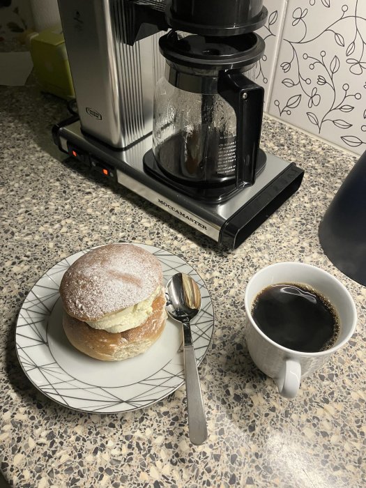 Kaffebryggare, semla på tallrik, svart kaffe i kopp, köksbänk.