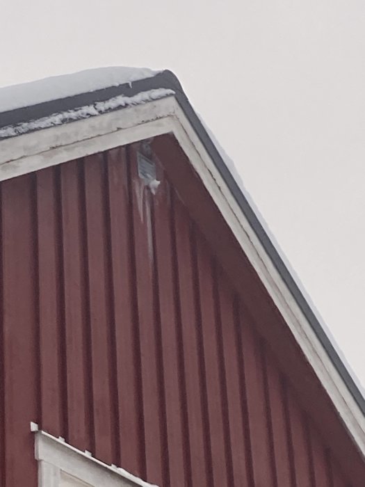 Rött hus med snötäckt takkant och istappar under en mulen himmel.