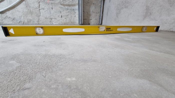 En gul vattenpass på grå betonggolv framför vit betongvägg. Byggverktyg för att kontrollera ytnivåer.