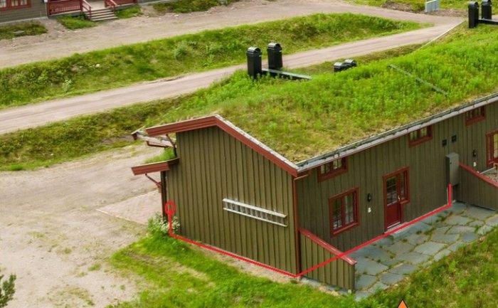 Grönt tak på byggnad, röda fönsterkarmar, omgiven av grusväg, markerade konturer med rött.