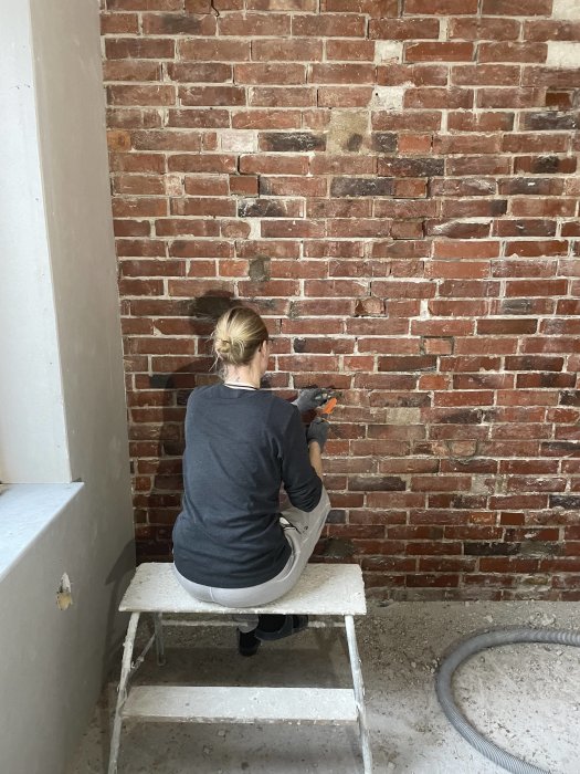 Person renoverar tegelvägg inomhus, sitter på en pall, håller en borr eller mejsel.