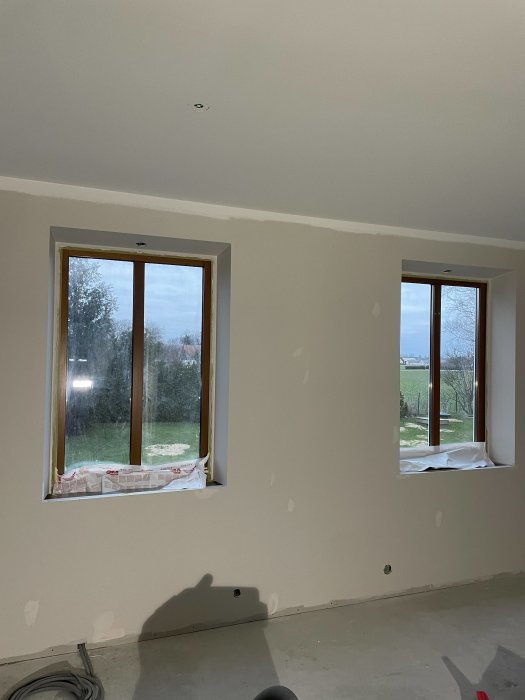 Omfärgat rum under renovering med två fönster, oskyddade golv och väggar i behov av ytterligare arbete.