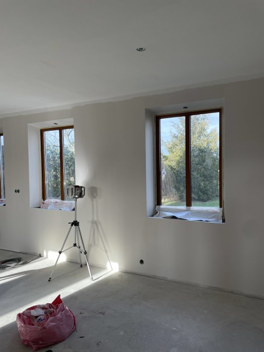 Ett omålade rum under renovering med tomma väggar, fönster, golv utan golvbeläggning och en arbetslampa på stativ.