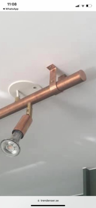 Vit skärm, kopparfärgad gardinstång med detaljer, spotlight, grå vägg, del av möbel under.
