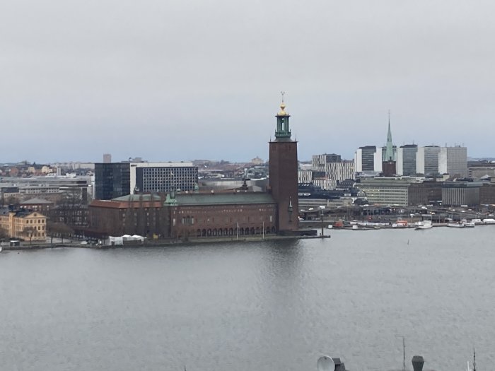 Stadssiluett med en framträdande byggnad nära vattnet, molnig himmel, båtar, modernt och historiskt arkitektursamspel.