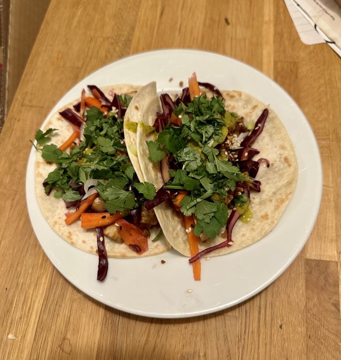 Två tacos med färska grönsaker och koriander toppade på mjuka tortillabröd, serverade på en vit tallrik.