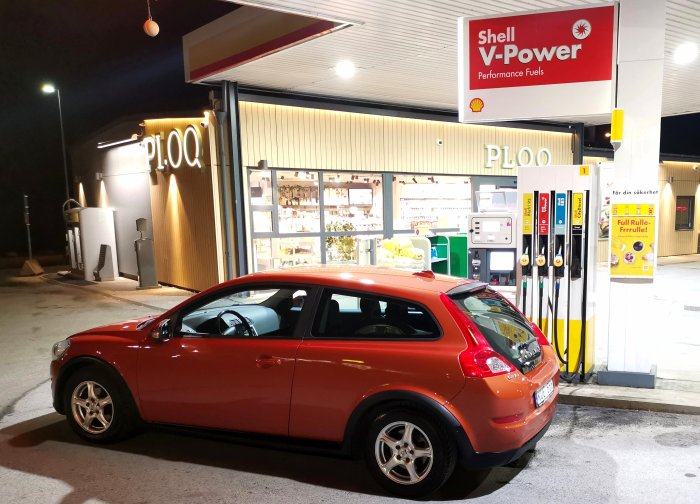 En röd bil står parkerad vid en Shell bensinstation nattetid, belysning på, ingen synlig aktivitet.