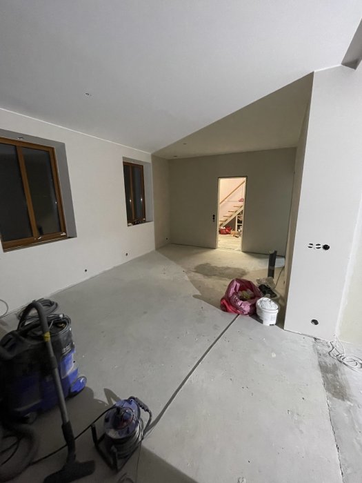 Ett tomt rum under renovering med synliga kablar, en dammsugare, fönster och en öppen dörr.