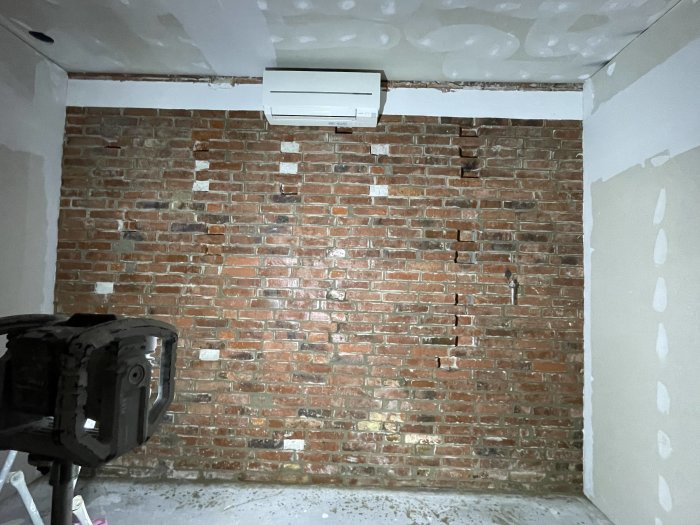Renoveringsarbete, röd tegelvägg, luftkonditioneringsenhet, ojämnt spacklade gipsväggar, delvis slutfört tak, byggnadsarbetsplats.