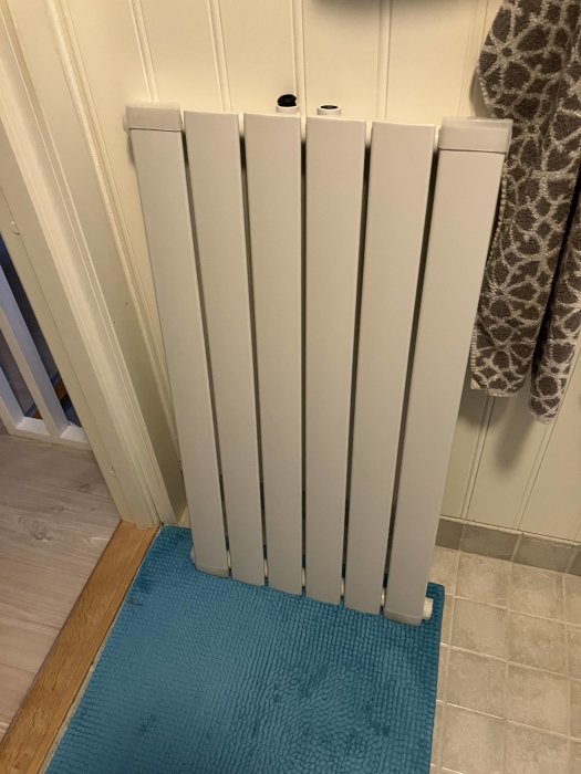 Vit radiator framför en veckad vägg, på en turkos badrumsmatta, bredvid en brun handduk.