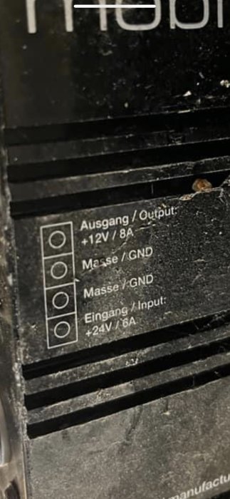 Etikett på elektronisk enhet med specifikationer: utgång 12V/8A, jord, ingång 24V/6A. Anslutningar synliga. Nedsmutsad.