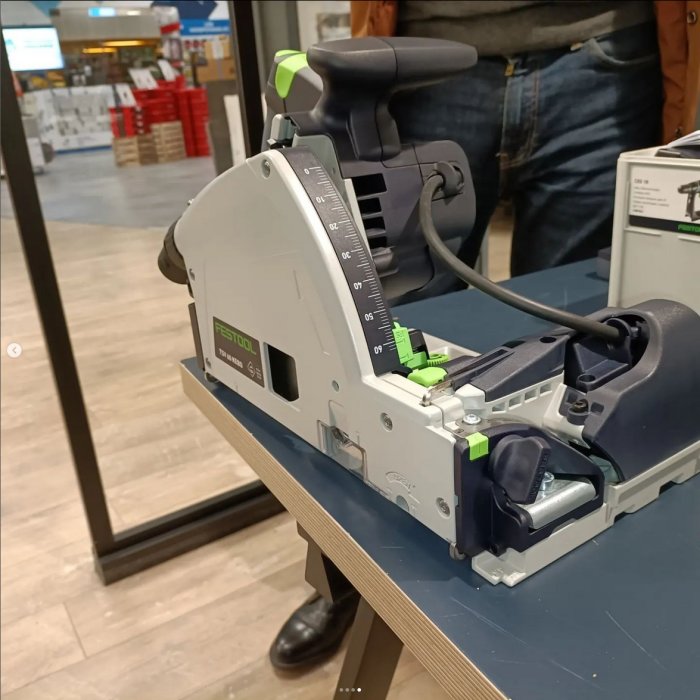 Festool cirkelsåg på bänk i butik, delvis synlig person i bakgrunden, arbetsredskap, ingen användning, inomhusmiljö.