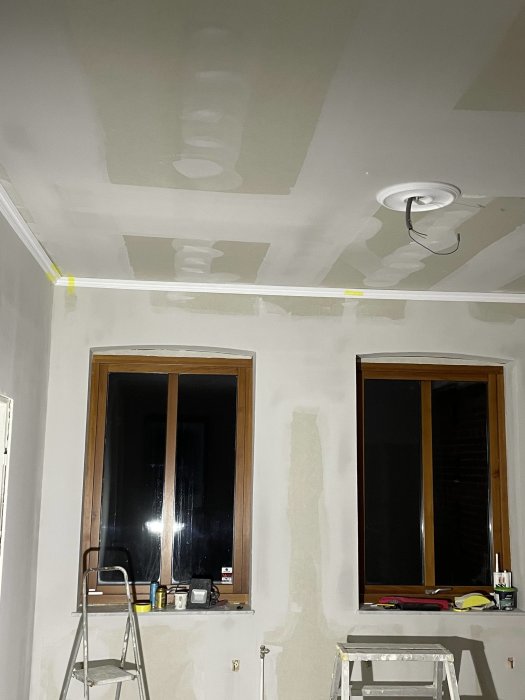 Ett rum under renovering med spacklade väggar, tejpade lister, en stege och fönster som speglar natten.